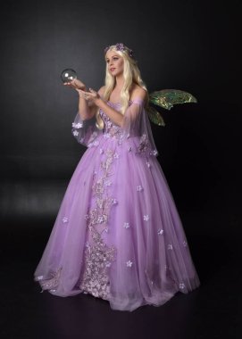 full length portrait of a blonde girl wearing a fantasy fairy inspired costume,  long purple ball gown with fairy wings,   sitting pose  on a dark studio background. clipart