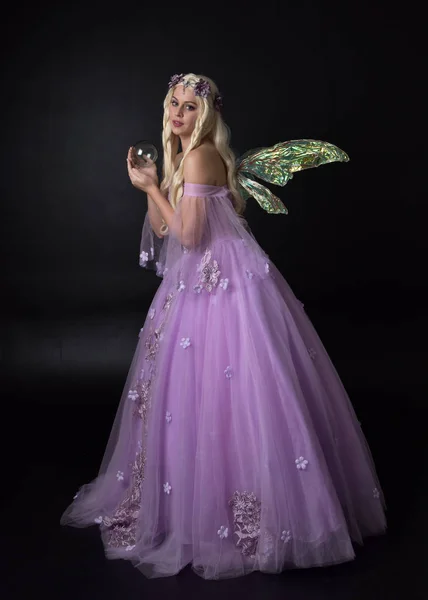 full length portrait of a blonde girl wearing a fantasy fairy inspired costume,  long purple ball gown with fairy wings,   sitting pose  on a dark studio background.