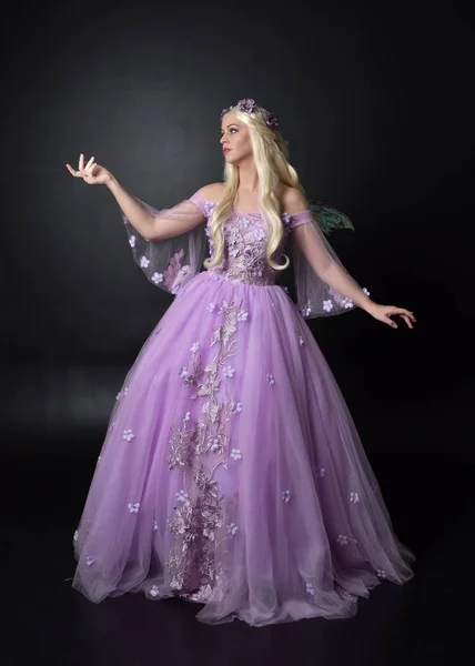 full length portrait of a blonde girl wearing a fantasy fairy inspired costume,  long purple ball gown with fairy wings,   sitting pose  on a dark studio background.