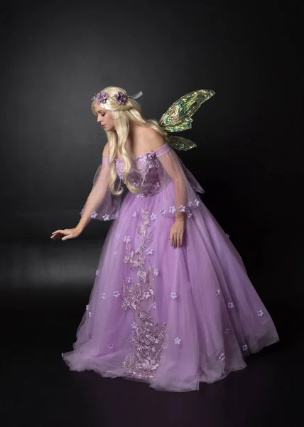 full length portrait of a blonde girl wearing a fantasy fairy inspired costume,  long purple ball gown with fairy wings,   sitting pose  on a dark studio background.