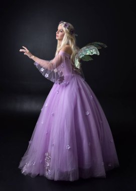 bir fantezi peri ilham kostüm giyen bir sarışın kız tam uzunlukta portre, peri kanatları ile uzun mor balo elbisesi, karanlık bir stüdyo arka planda ayakta poz.