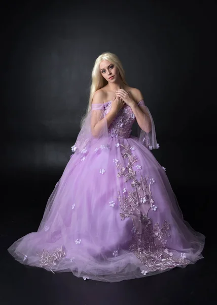full length portrait of a blonde girl wearing a fantasy fairy inspired costume,  long purple ball gown with fairy wings,   sitting pose on a dark studio background.