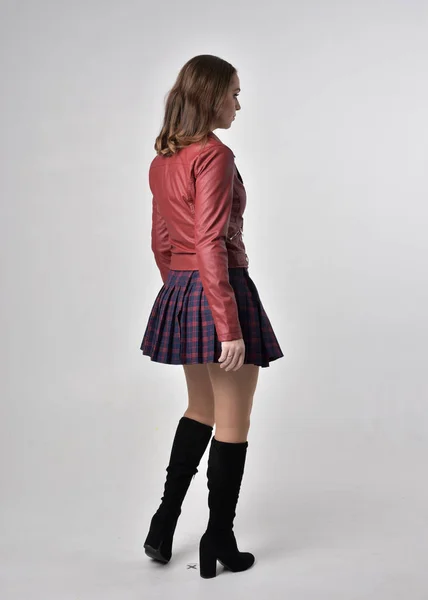赤い革のジャケットとチェック柄のスカートを着たブルネットの女の子の完全な長さの肖像画 クリームスタジオの背景にカメラに背中を持って立っているポーズ — ストック写真