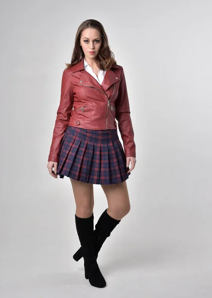 赤い革のジャケットとチェック柄のスカートを着たブルネットの女の子のフルレングスの肖像画 クリームスタジオの背景に立ってポーズ — ストック写真