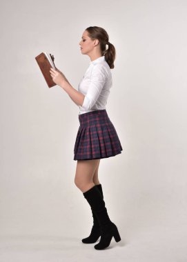 beyaz gömlek ve ekose etek giyen bir esmer kız tam uzunlukta portre, ayakta poz ve bir krem stüdyo arka plan karşı bir kitap tutarak.