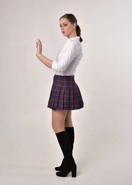 一个黑发女孩穿着白色衬衫和格子裙制服的全长肖像 站在奶油工作室背景的侧轮廓姿势 — 图库照片