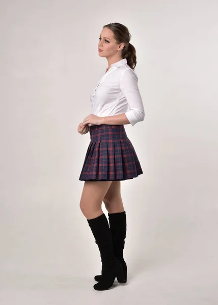 白いシャツとチェック柄のスカートのユニフォームを着たブルネットの女の子のフルレングスの肖像画 クリームスタジオの背景に対してサイドプロファイルで立っているポーズ — ストック写真