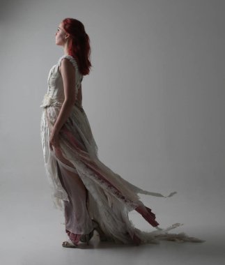 yırtık ve yırtık gelinlik giyen kırmızı saçlı kız tam uzunlukta portre. Kontrastlı gölge aydınlatması ile stüdyo arka planına karşı ayakta poz.