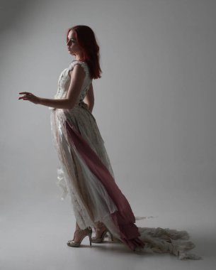 full length portrait of red haired girl wearing torn and tattered wedding dress. Standing pose against a studio background with contrasty shadow lighting. clipart