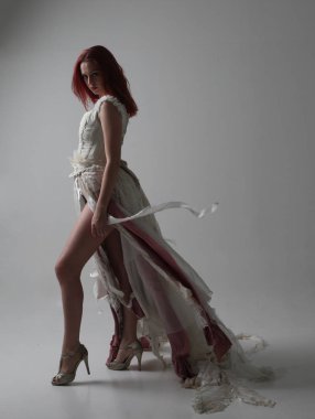 full length portrait of red haired girl wearing torn and tattered wedding dress. Standing pose against a studio background with contrasty shadow lighting. clipart