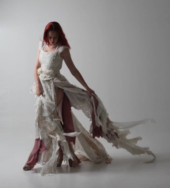 yırtık ve yırtık gelinlik giyen kırmızı saçlı kız tam uzunlukta portre. Kontrastlı gölge aydınlatması ile stüdyo arka planına karşı ayakta poz.