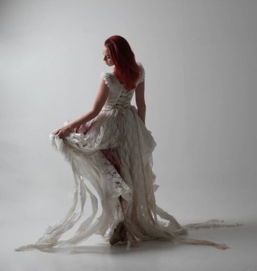 yırtık ve yırtık gelinlik giyen kırmızı saçlı kız tam uzunlukta portre. Kontrastlı gölge aydınlatması ile stüdyo arka planına karşı ayakta poz.