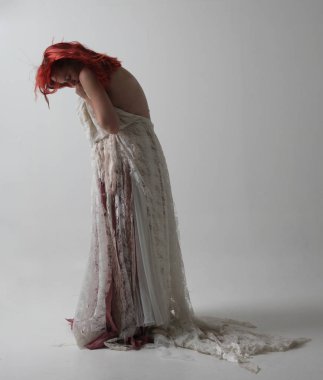 yırtık ve yırtık gelinlik giyen kırmızı saçlı kız tam uzunlukta portre. Kontrastlı gölge aydınlatması ile stüdyo arka planına karşı ayakta poz.