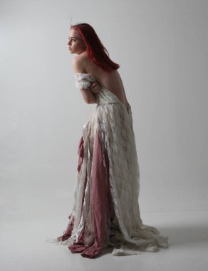 yırtık ve yırtık gelinlik giyen kırmızı saçlı kız tam uzunlukta portre. Kontrastlı gölge aydınlatması ile stüdyo arka planına karşı ayakta poz.