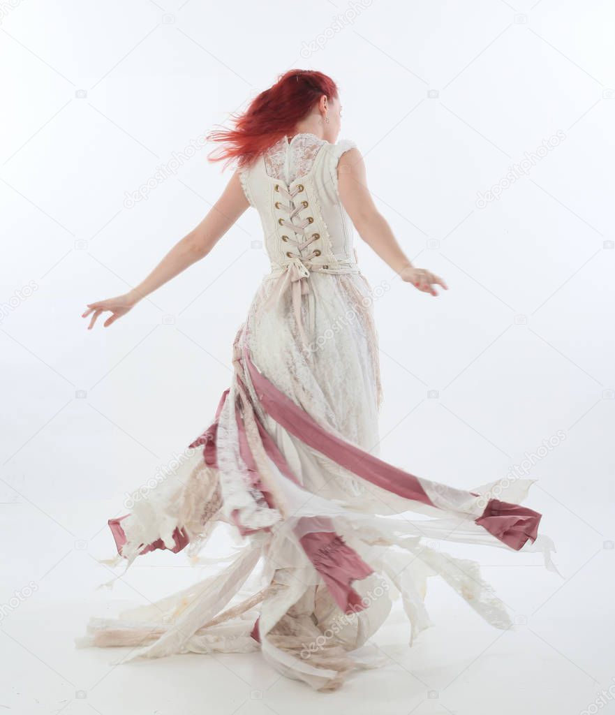 full length portrait of red haired girl wearing torn and tattered wedding dress. Standing pose against a  white studio background.