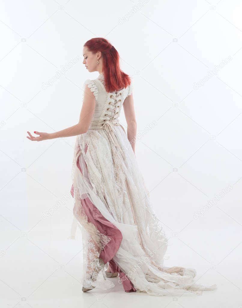 full length portrait of red haired girl wearing torn and tattered wedding dress. Standing pose against a  white studio background.
