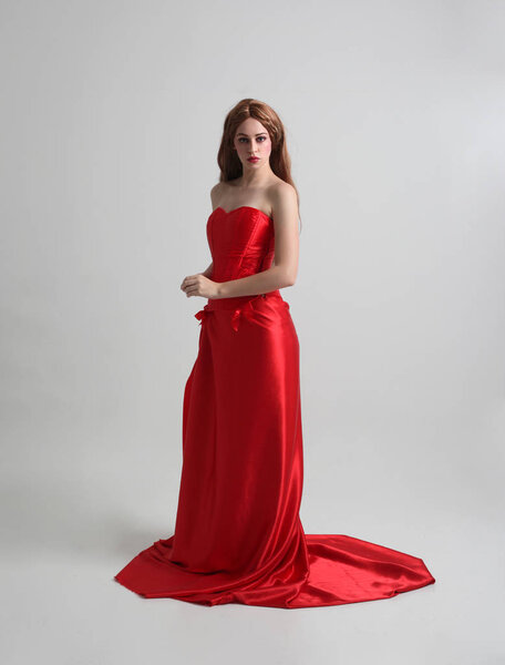 портрет девушки в длинном красном шелковом платье, стоящей в позе на сером фоне студии
.