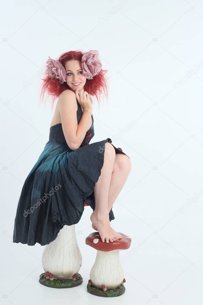 full length portrait of red haired girl wearing fairy costume, seated pose on a white studio background with mushroom props.
