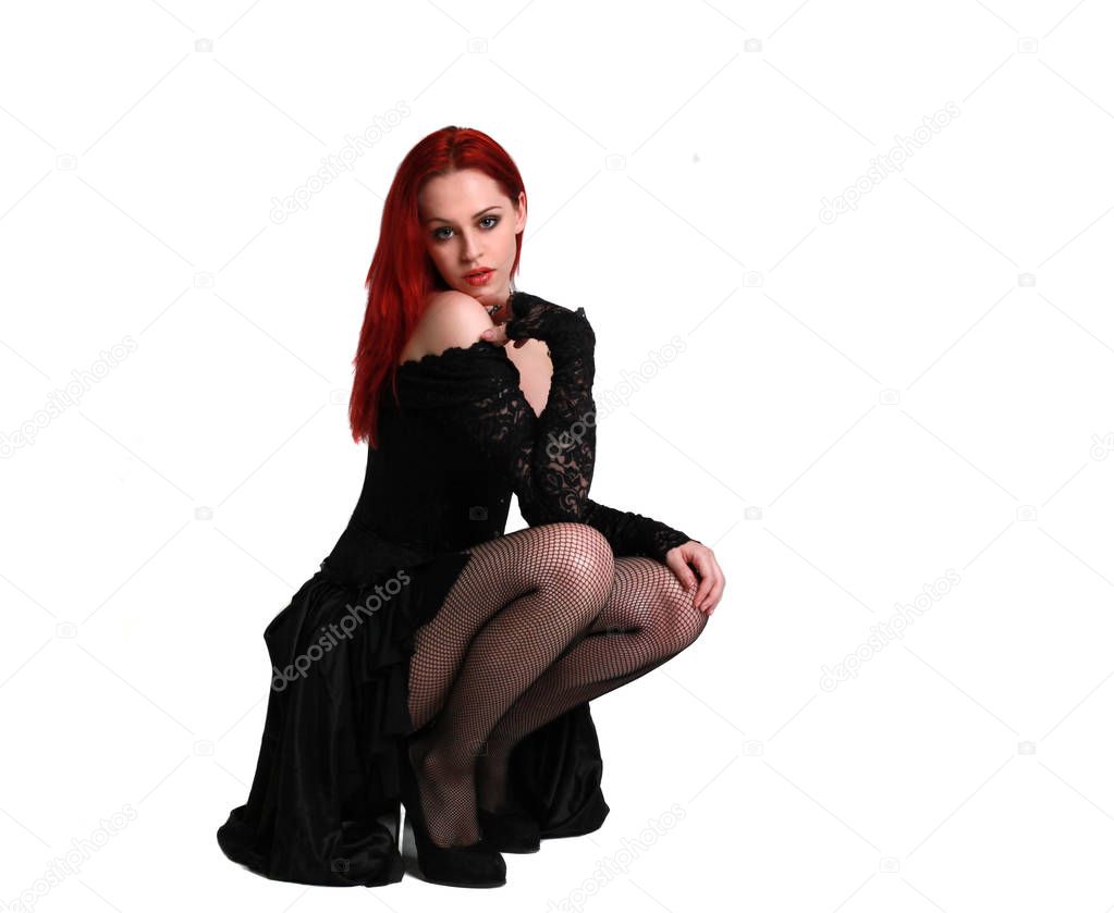 full length portrait of a  red haired girl wearing a  black gothic gown, Seated pose on a grey studio background.