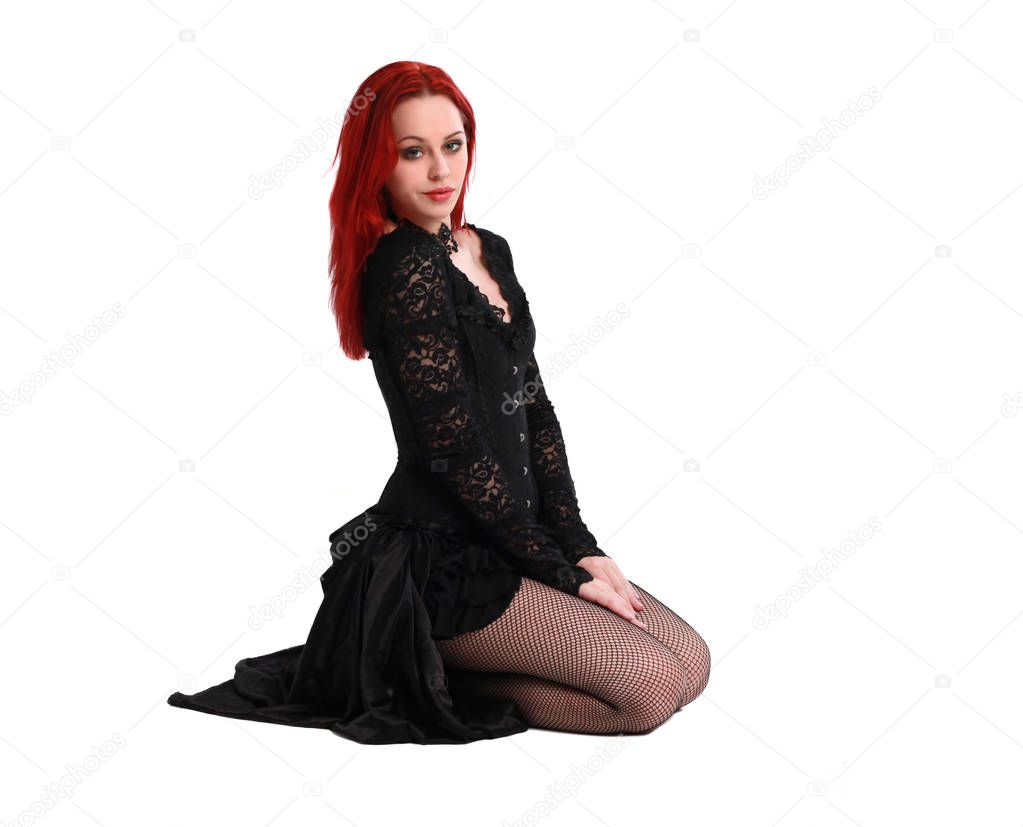full length portrait of a  red haired girl wearing a  black gothic gown, Seated pose on a grey studio background.