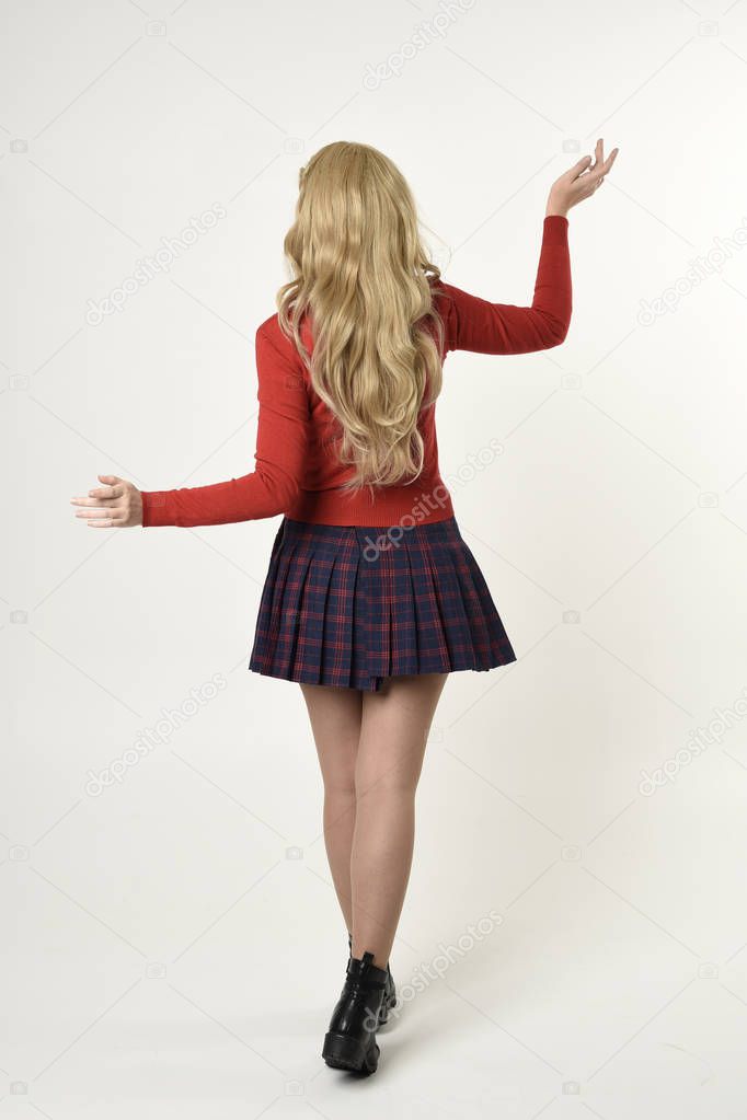 full length portrait of blonde girl wearing red cardigan with tie and plaid skirt, school uniform, standing pose facing away from the camera, on a white studio background.