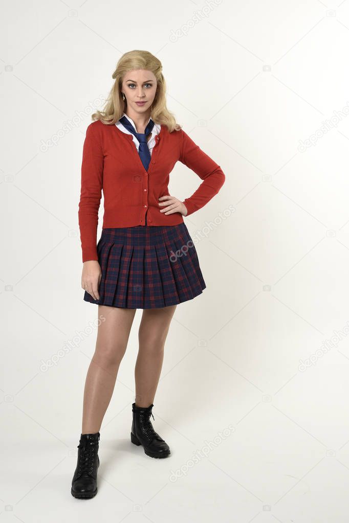 full length portrait of blonde girl wearing red cardigan with tie and plaid skirt, school uniform, standing,  walking  pose facing the camera, on a white studio background.