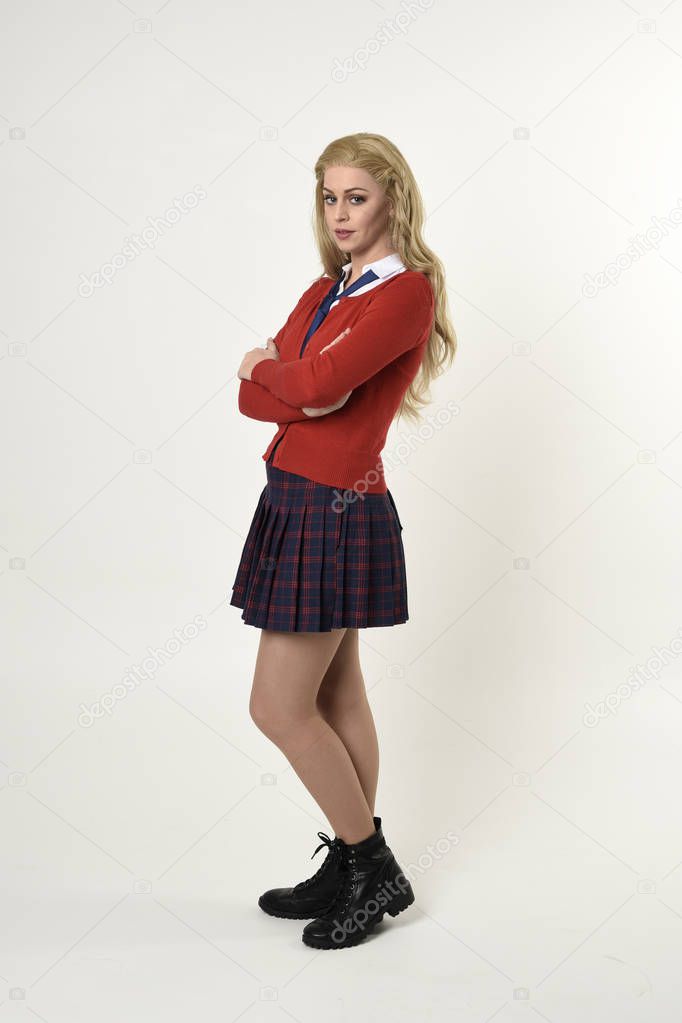 full length portrait of blonde girl wearing red cardigan with tie and plaid skirt, school uniform, standing,  walking  pose facing the camera, on a white studio background.