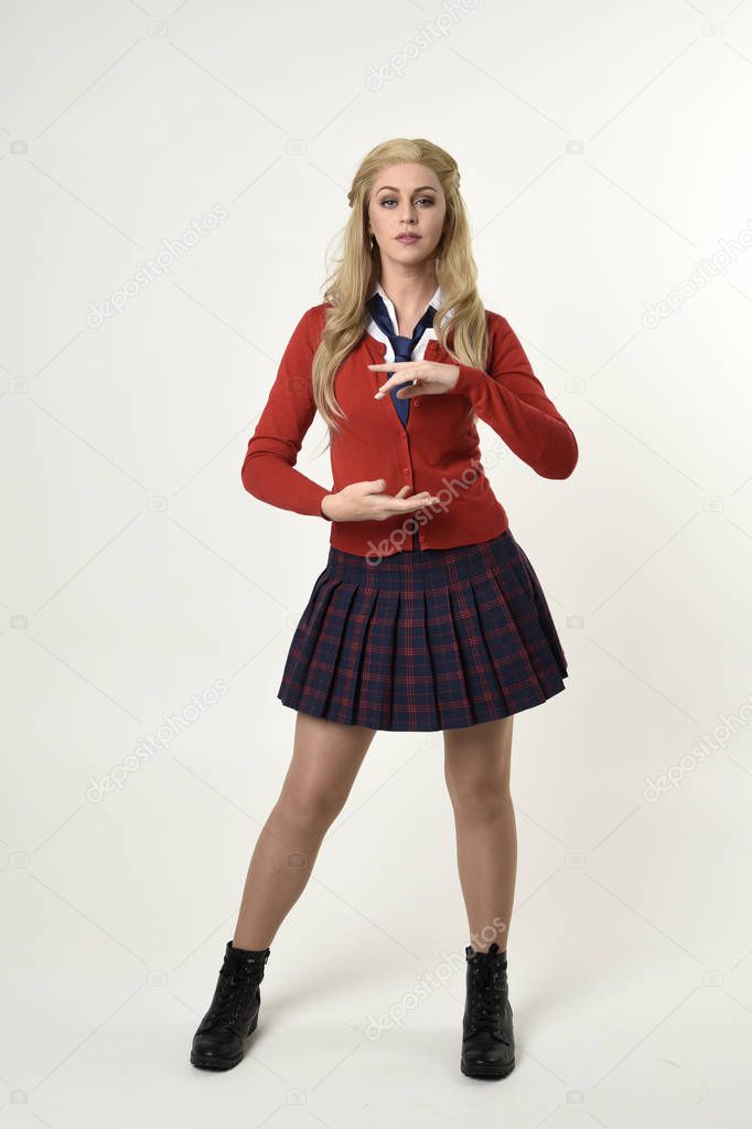 full length portrait of blonde girl wearing red cardigan with tie and plaid skirt, school uniform, standing,  walking  pose facing the camera, on a white studio background.