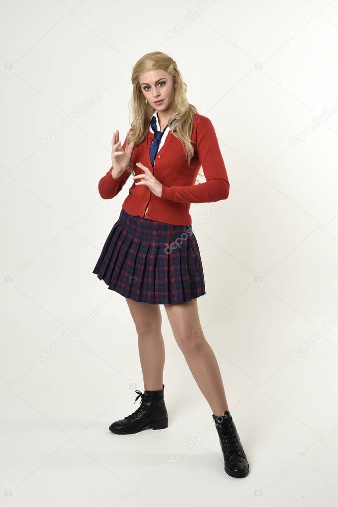 full length portrait of blonde girl wearing red cardigan with tie and plaid skirt, school uniform, standing,  walking  pose facing the camera, on a white studio background.