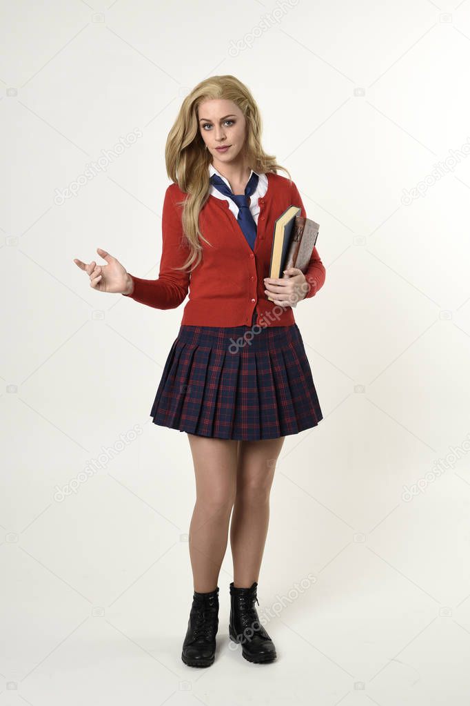 full length portrait of blonde girl wearing red cardigan with tie and plaid skirt, school uniform, standing  pose holding books,  facing the camera, on a white studio background.