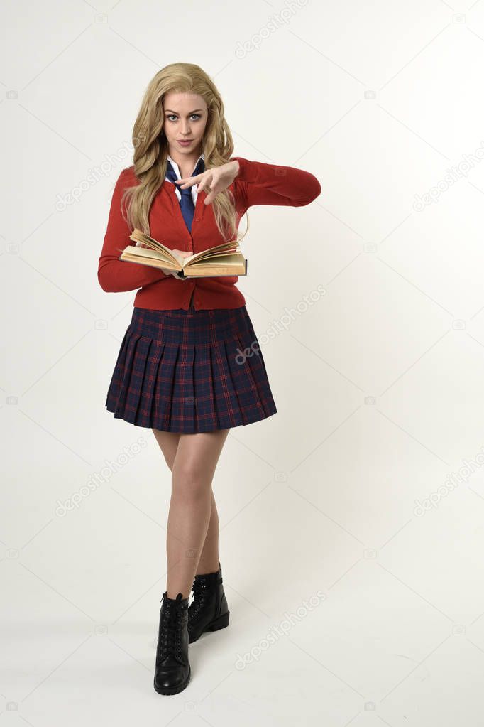 full length portrait of blonde girl wearing red cardigan with tie and plaid skirt, school uniform, standing  pose holding books,  facing the camera, on a white studio background.
