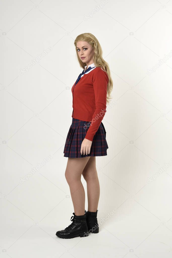 full length portrait of blonde girl wearing red cardigan with tie and plaid skirt, school uniform, side profile, standing  pose, on a white studio background.