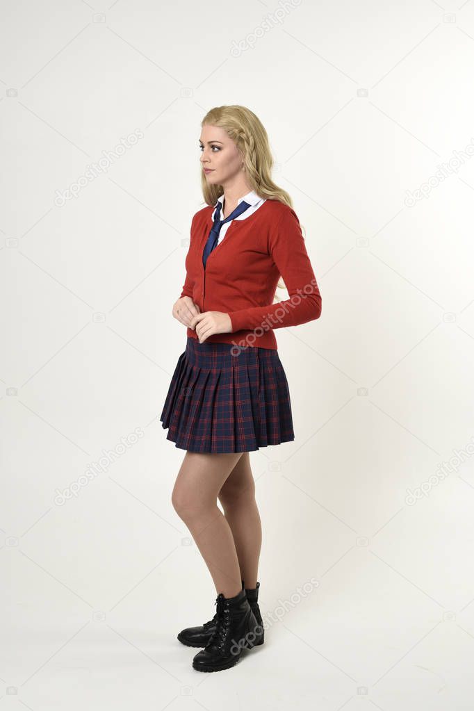 full length portrait of blonde girl wearing red cardigan with tie and plaid skirt, school uniform, side profile, standing  pose, on a white studio background.