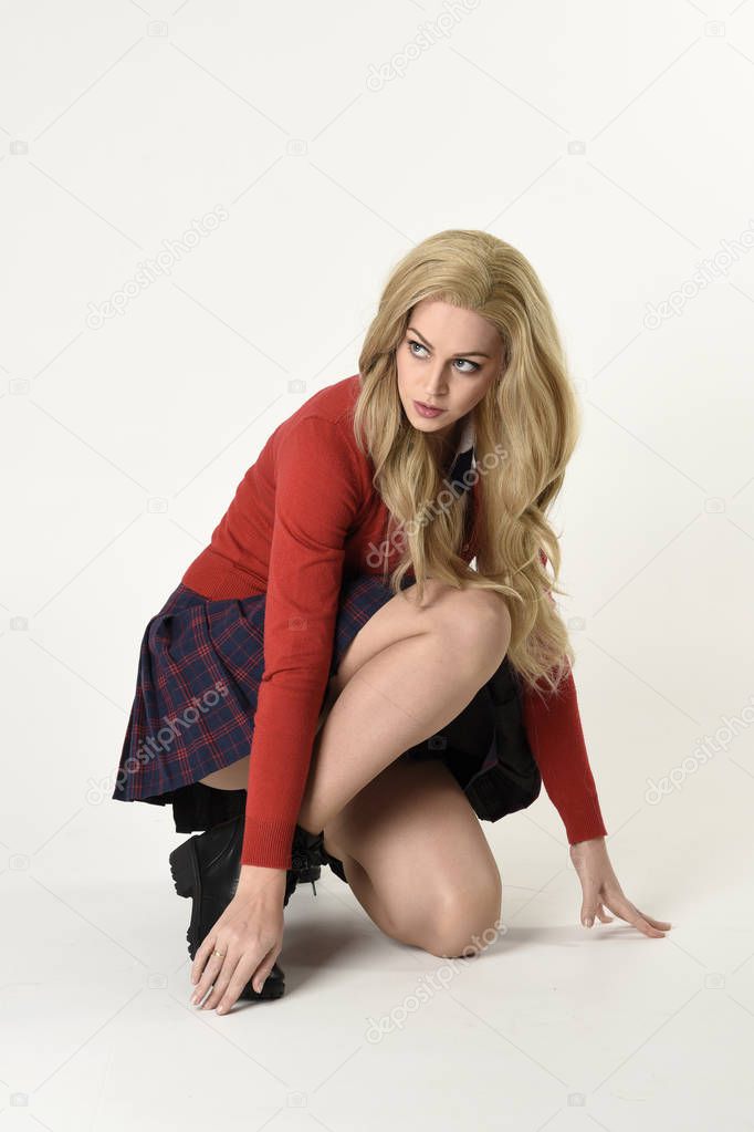 full length portrait of blonde girl wearing red cardigan with tie and plaid skirt, school uniform. Sitting on the ground with  a white studio background.