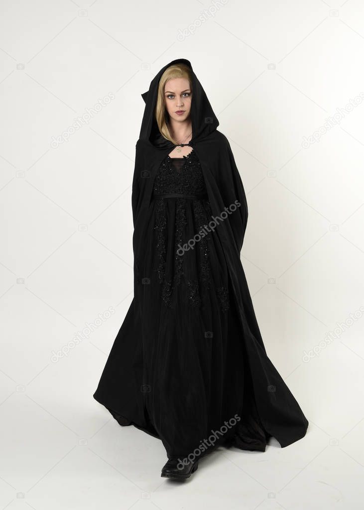 full length portrait of blonde girl wearing long black flowing cloak, standing pose  with  a white studio background.