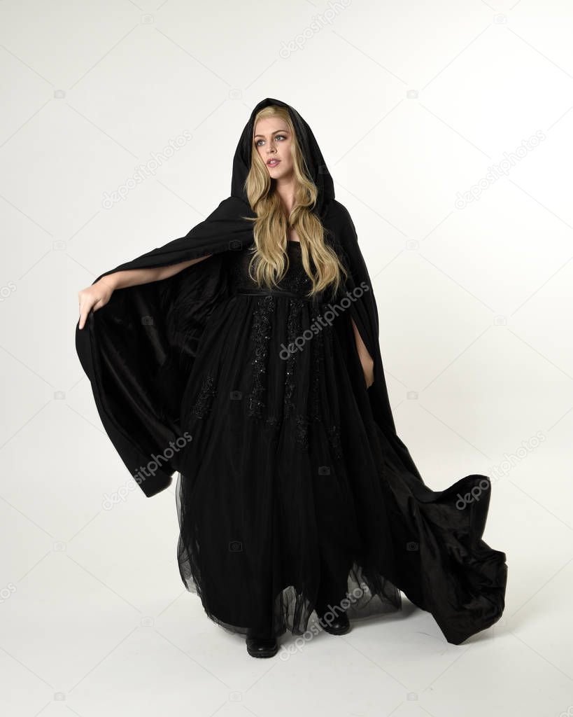 full length portrait of blonde girl wearing long black flowing cloak, standing pose  with  a white studio background.
