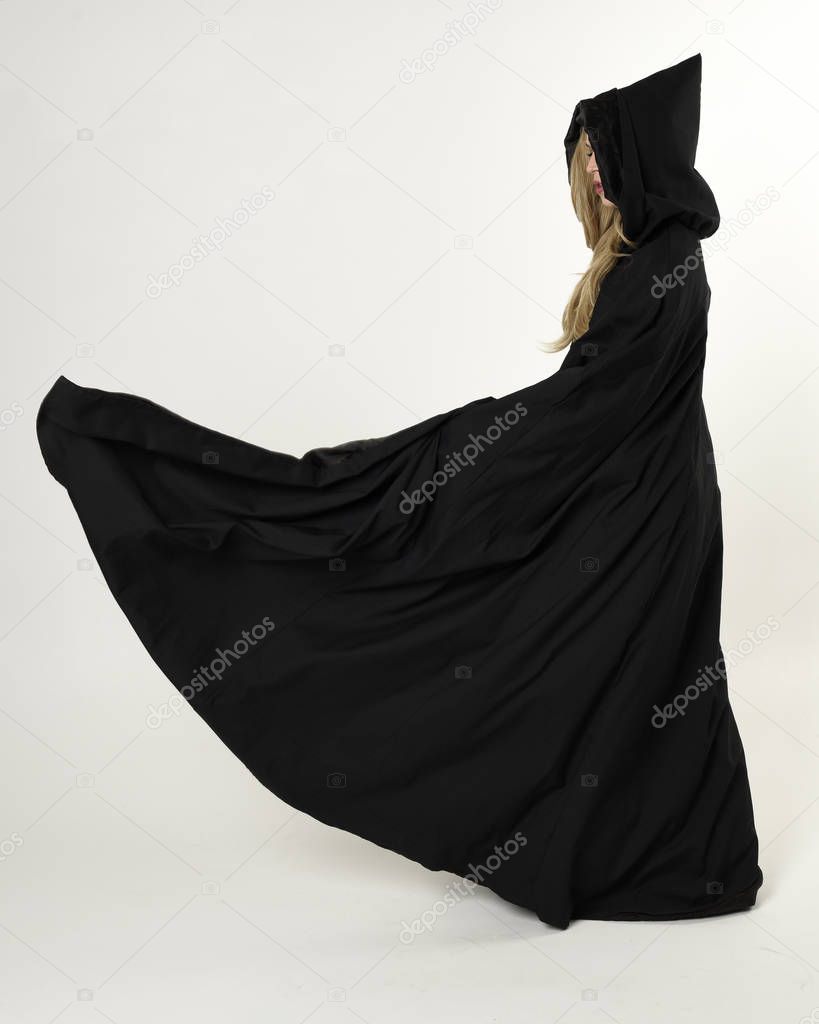 full length portrait of blonde girl wearing long black flowing cloak, standing pose  with  a white studio background.