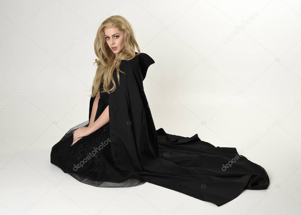 full length portrait of blonde girl wearing long black flowing cloak, sitting on the floor  with  a white studio background.