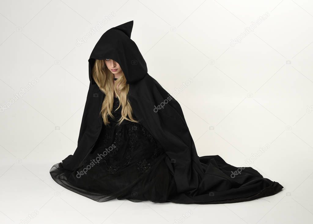 full length portrait of blonde girl wearing long black flowing cloak, sitting on the floor  with  a white studio background.