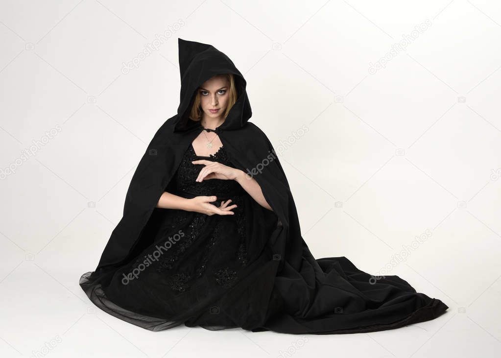 full length portrait of blonde girl wearing long black flowing cloak, sitting on the floor  with  a white studio background.