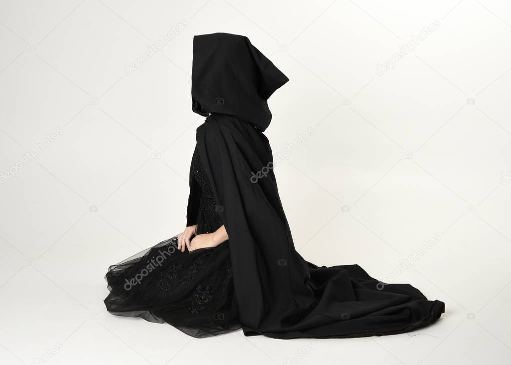 full length portrait of blonde girl wearing long black flowing cloak, sitting on the floor  with  a white studio background.