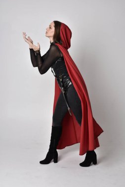 Uzun boylu, esmer, siyah deri fantezi kostümlü, uzun kırmızı süper kahraman pelerinli bir kadının portresi. Stüdyo arka planında ayakta durma.