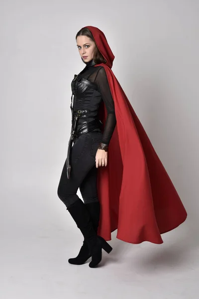 長い赤いスーパーヒーローのケープと黒革のファンタジーの衣装を身に着けているかなりブルネットの女性の完全な長さの肖像画 スタジオの背景に立ちポーズをとって — ストック写真