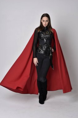 full length portrait of a pretty brunette woman wearing black leather fantasy costume with long red superhero cape. standing pose on a studio background. clipart