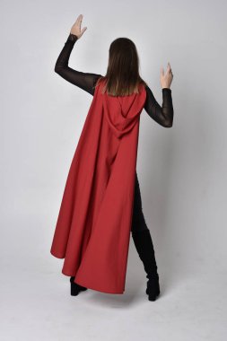 Uzun boylu, esmer, siyah deri fantezi kostümlü, uzun kırmızı süper kahraman pelerinli bir kadının portresi. Stüdyo arka planında ayakta durma.