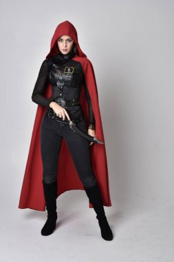 full length portrait of a pretty brunette woman wearing black leather fantasy costume with long red superhero cape. standing pose on a studio background. clipart