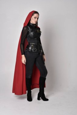 Uzun boylu, esmer, siyah deri fantezi kostümlü, uzun kırmızı süper kahraman pelerinli bir kadının portresi. Stüdyo arka planında ayakta durma.