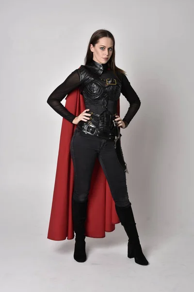 full length portrait of a pretty brunette woman wearing black leather fantasy costume with long red superhero cape. standing pose on a studio background.