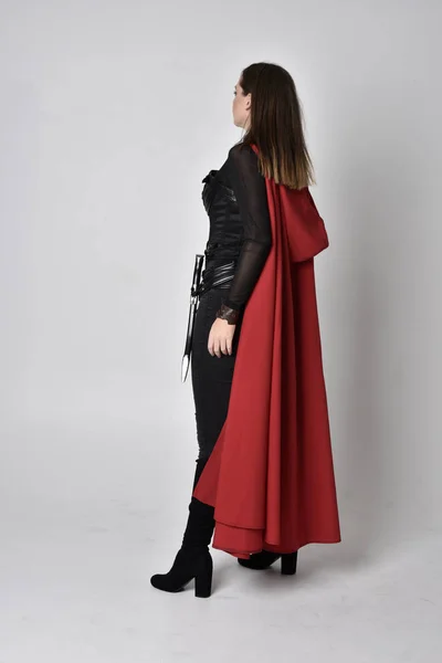 全长肖像一个漂亮的黑发女人穿着黑色皮革幻想服装与长长的红色超级英雄斗篷 站在工作室背景上的姿势 — 图库照片