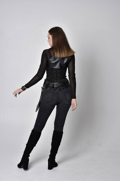 full length portrait of a pretty brunette woman wearing black leather fantasy costume with a dagger. standing pose facing away from camera on a studio background.